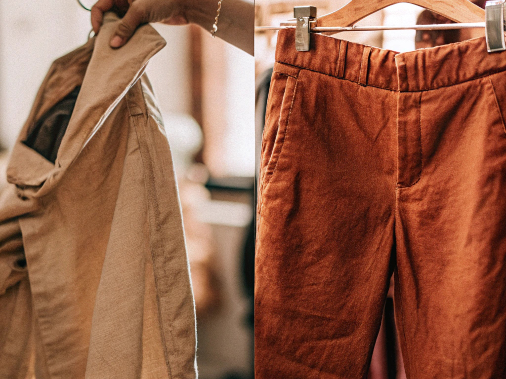 Hand holding beige linen pants and a hanging pair of rust-colored linen trousers, emphasizing their texture and tailored design.
