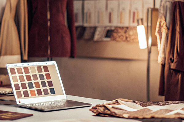 A designer’s workspace with a laptop displaying color swatches, clothing sketches on a board, and mannequins wearing elegant outfits.