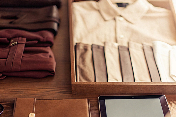 A wooden table with neatly arranged clothing, accessories, a leather notebook, an iPad, and a tray of folded neutral-toned shirts.