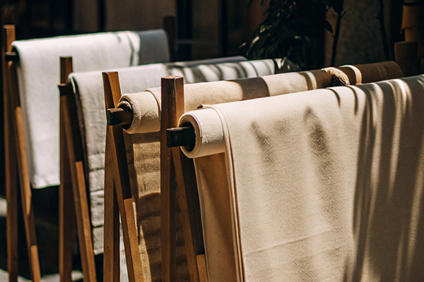Rolls of natural beige fabric drying on wooden stands under dappled sunlight outdoors.