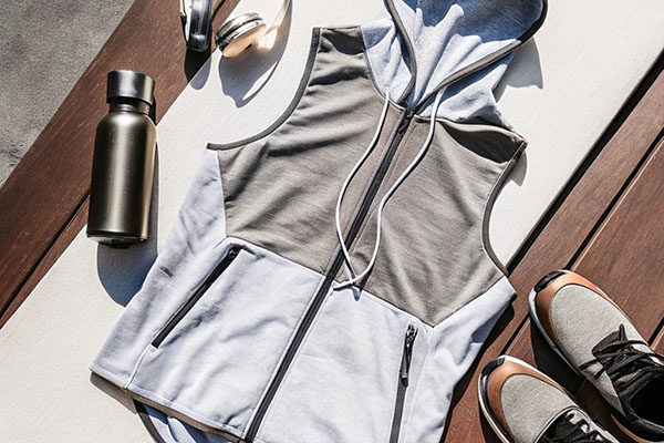 Gray and white sleeveless hoodie with a zipper, placed alongside a water bottle, headphones, and athletic shoes on a wooden surface.