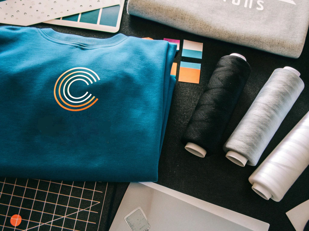 A blue hoodie with a circular logo, alongside thread spools, fabric samples, and design tools on a creative workspace.