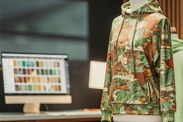 Mannequin wearing a camouflage-patterned hoodie with a computer displaying color palettes in the background.