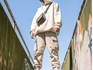 Person wearing a cream hoodie, cargo pants, and a crossbody bag standing outdoors against a clear blue sky.