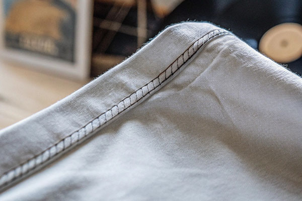 Detailed view of a light-colored t-shirt seam showcasing precise stitching and soft fabric texture with a blurred vinyl record in the background.