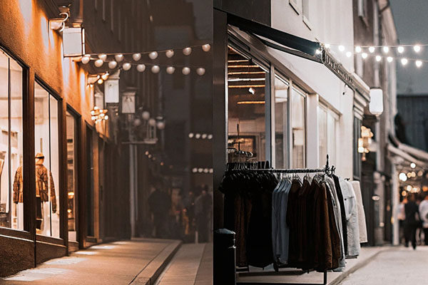 A night-time view of a clothing store with illuminated windows, garments displayed outside on racks, and string lights decorating the street.