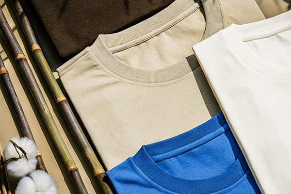 A collection of beige, brown, blue, and white cotton t-shirts laid flat beside bamboo sticks and fluffy raw cotton branches.