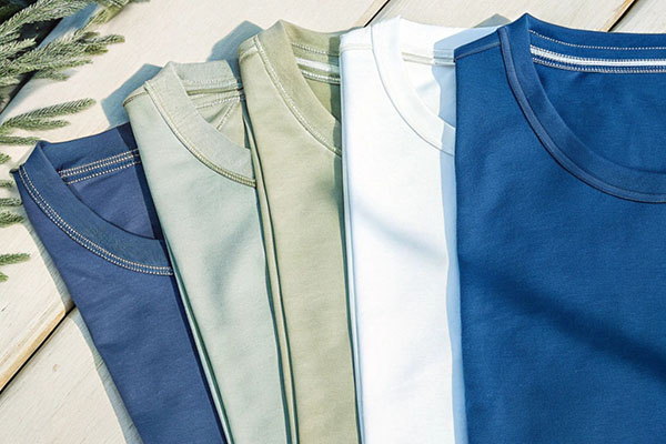 A row of five neatly folded t-shirts in blue, green, white, and olive shades, displayed on a light wooden surface with soft greenery in the background.
