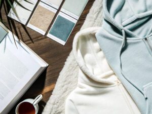 Light blue and white hoodies paired with fabric swatches, a book, and a cup of tea on a wooden table, evoking comfort and style.