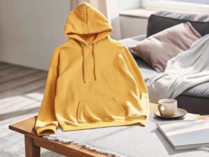 Yellow hoodie displayed on a wooden table in a cozy living room setting, with a cup of coffee and soft cushions in the background.