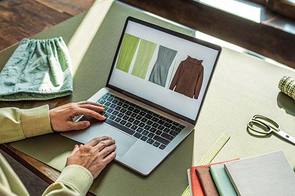 Person in light green sleeve viewing garment designs and fabric textures on a laptop screen, with sewing tools on the table.