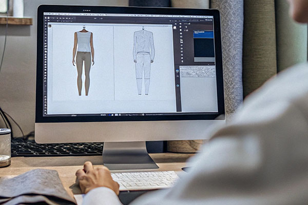 A designer working on a computer displaying front and back views of clothing sketches, with fabric rolls in the background.
