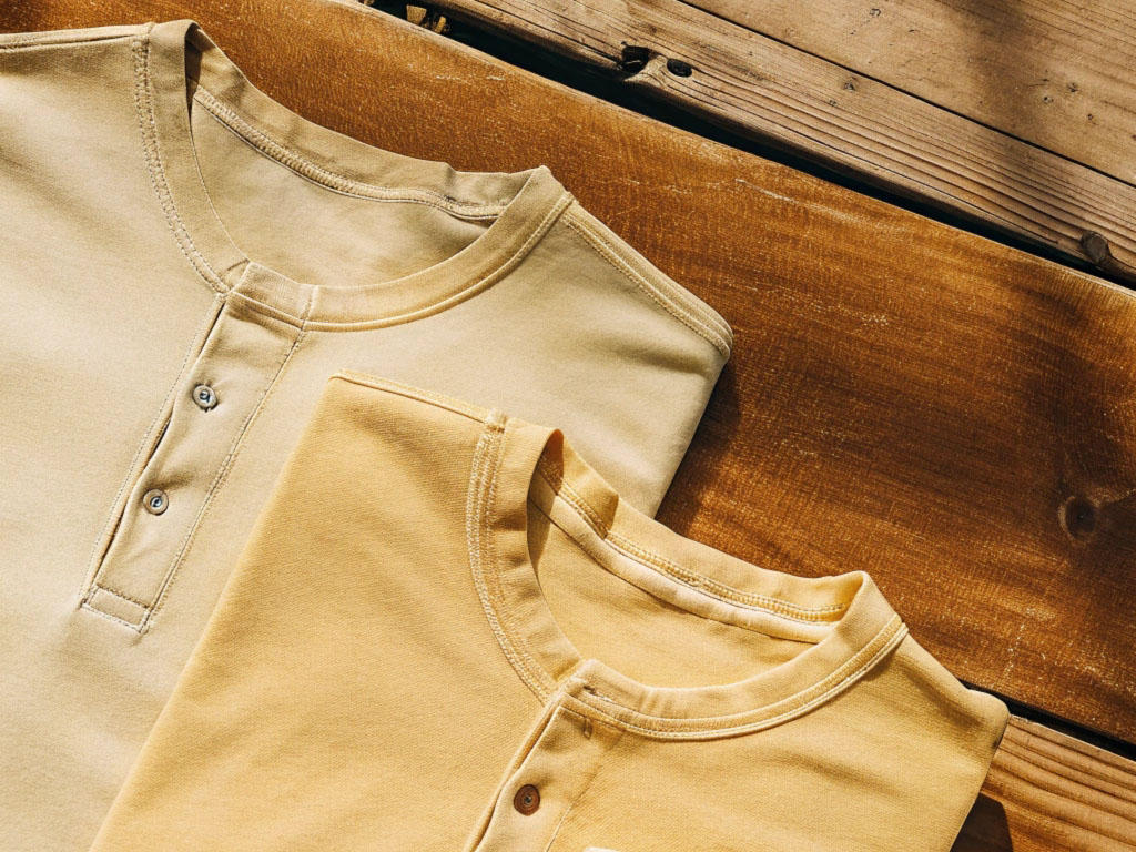 Three neatly folded earth-tone t-shirts in beige and mustard yellow shades, laid on a rustic wooden surface under sunlight.