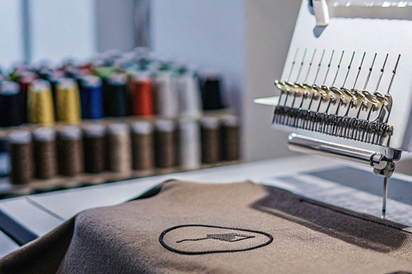 Close-up of an embroidery machine stitching a design onto fabric, with colorful thread spools blurred in the background.