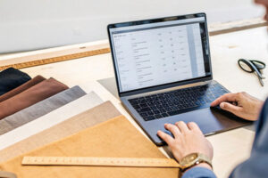 Person working on a laptop displaying a spreadsheet, with fabric swatches, a ruler, and scissors on a table.