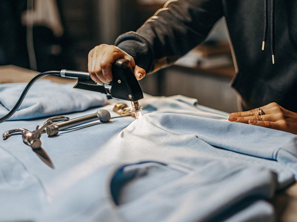 Person using a specialized tool to add detailing to a light blue fabric, with metal tools placed on the table.