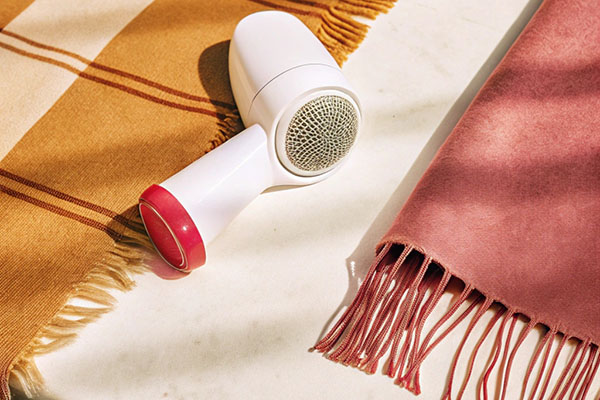 A white lint remover with a red handle placed between a mustard striped and a pink fringed fabric on a light surface.
