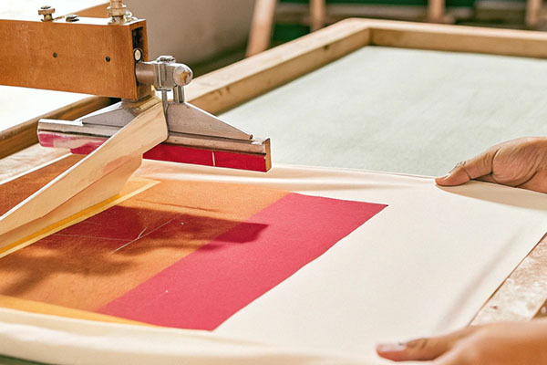Close-up of a screen printing machine applying bold red and orange ink onto white fabric, with hands holding the material steady in a workshop.