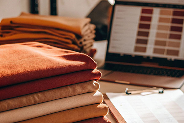 Neatly folded orange, tan, and beige fabrics stacked on a table with a laptop displaying color palettes in the background.