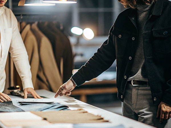 Two men examining fabric swatches and clothing designs on a well-lit table in a workspace, with coats hanging in the background.