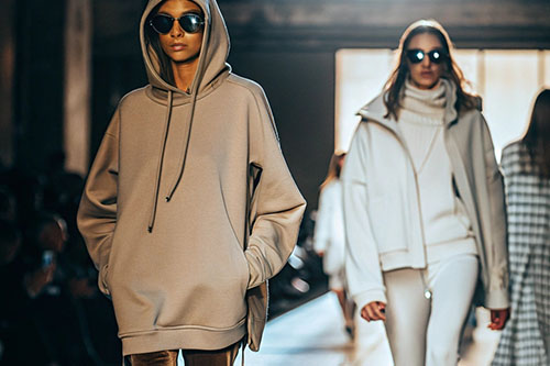 Models wearing stylish hoodies and winter outfits walking down a runway during a fashion show, with dramatic lighting.
