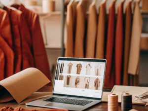 Laptop displaying clothing designs on a desk surrounded by fabric rolls, spools, and garments in warm tones.