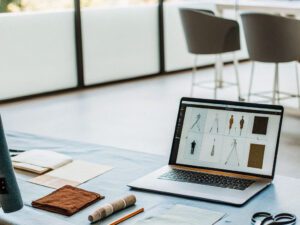 Open laptop displaying fashion sketches on a table with fabric samples, scissors, and pencils, in a bright, modern space.