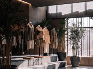 Mannequins dressed in earthy-toned outfits displayed in a minimalist showroom with large plants and natural lighting.