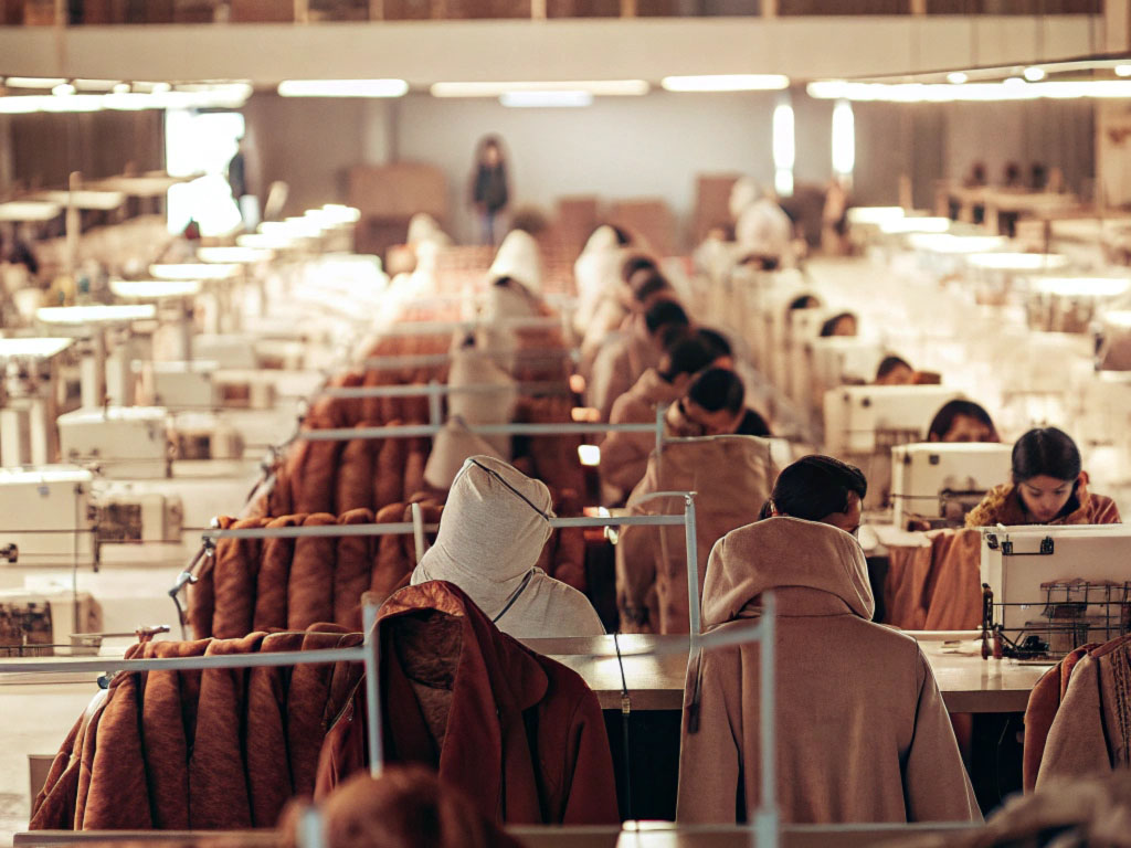 Workers in a large garment factory assembling clothing at sewing stations, surrounded by coats and machinery under bright industrial lighting.