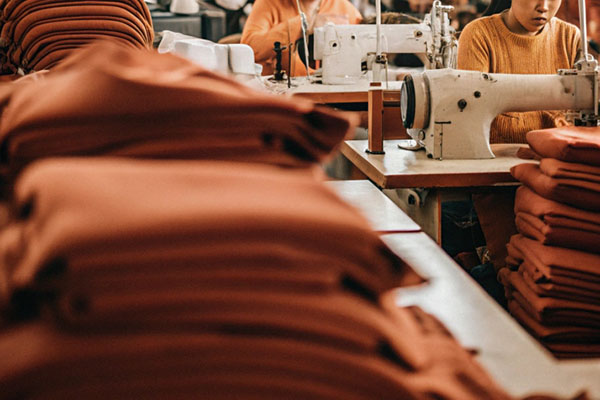 Workers sewing garments in a busy factory with stacks of orange fabric on tables.