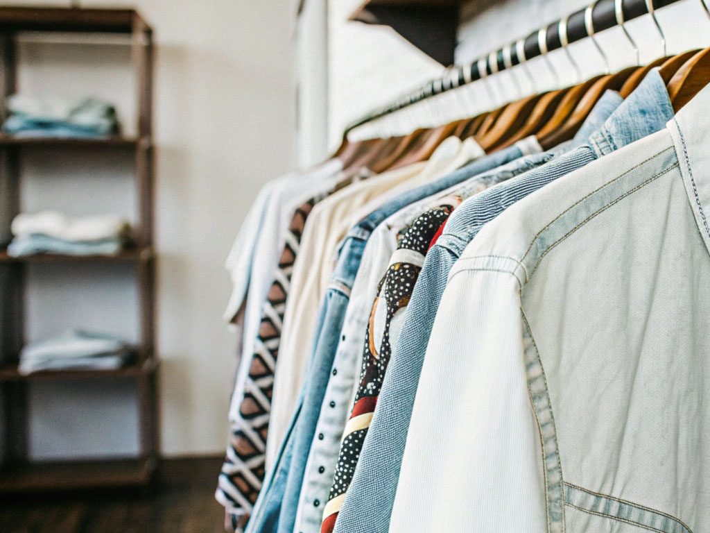 A row of denim jackets, shirts, and patterned clothing hanging on wooden hangers in a boutique with wooden shelving in the background.
