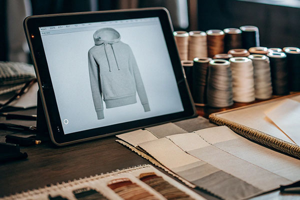 Tablet showing a hoodie sketch surrounded by fabric swatches, thread spools, and material samples on a workspace.