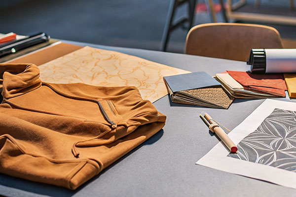 A mustard-colored hoodie on a table with fabric swatches, sketches, and design tools, showcasing a creative workspace.