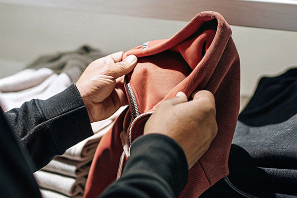Hands closely examining the fabric and quality of a red hoodie, with neatly folded clothes in the background.