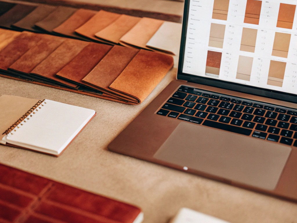 Laptop displaying leather samples alongside swatches, notebooks, and fabric in warm brown tones.