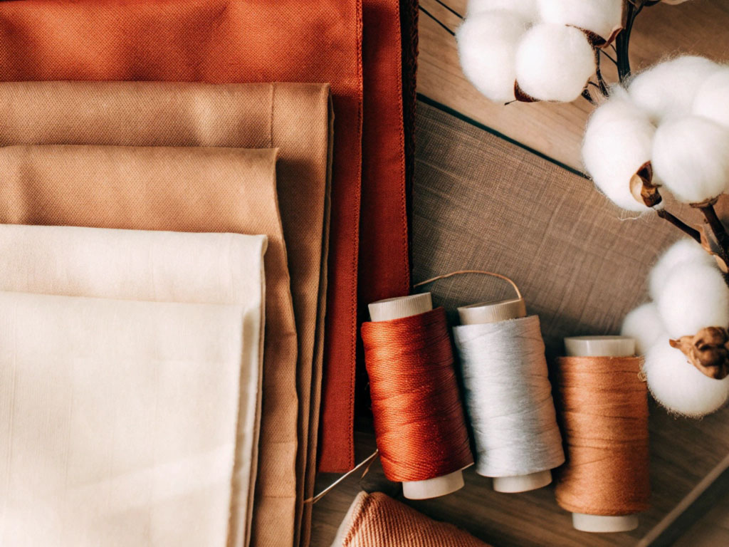 Assorted beige, brown, and orange fabrics, cotton spools, and raw cotton branches arranged on a wooden surface.