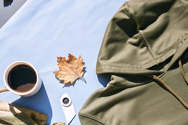 Olive green hoodie displayed alongside a cup of coffee, a dried leaf, and a light blue background.