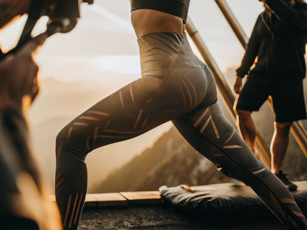 Woman wearing patterned workout leggings stretching during an outdoor exercise session at sunrise, with mountains in the background.