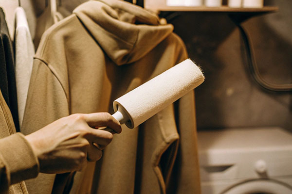 Hand using a lint roller on a beige hoodie hanging in a laundry room with a washer in the background.