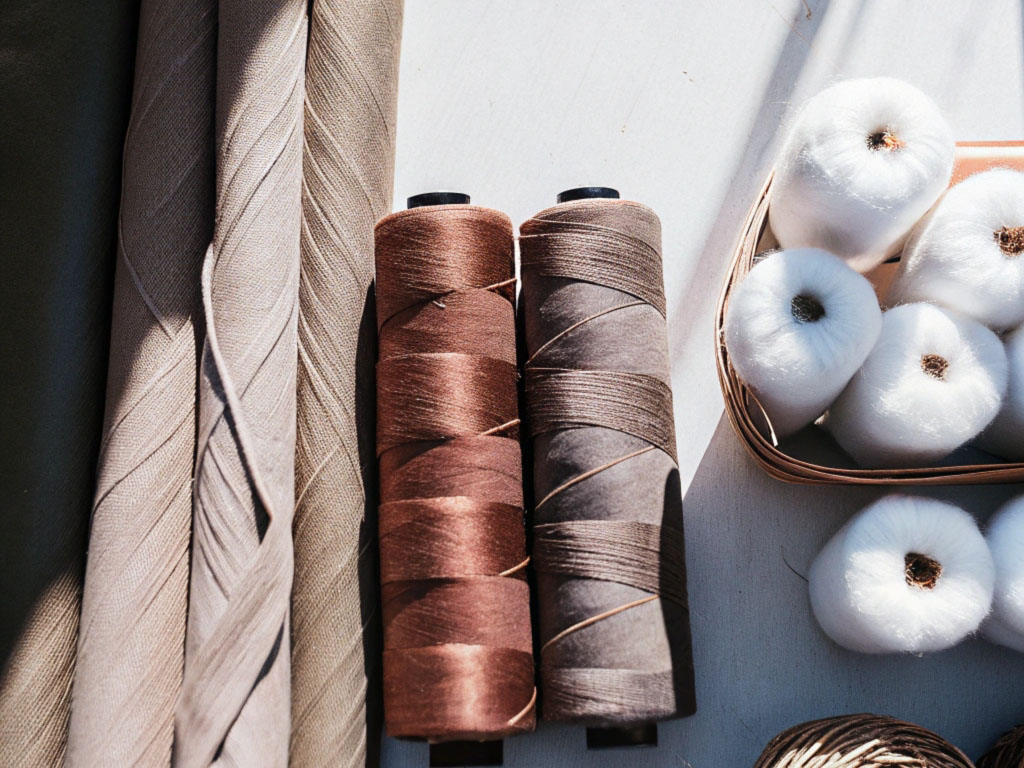 Brown and gray spools of thread alongside fabric rolls and white cotton balls on a table under sunlight.