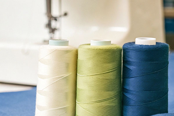Three spools of thread in cream, green, and blue shades placed on a blue fabric with a sewing machine in the background.