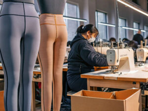 Seamstress working on a sewing machine in a factory setting with mannequins displaying leggings and fabric materials nearby.