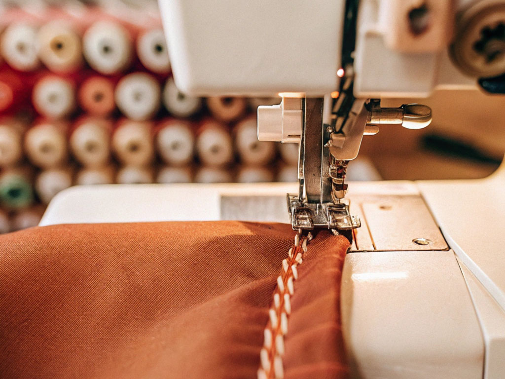 Close-up of a sewing machine creating a decorative stitch on rust-colored fabric with thread spools in the background.