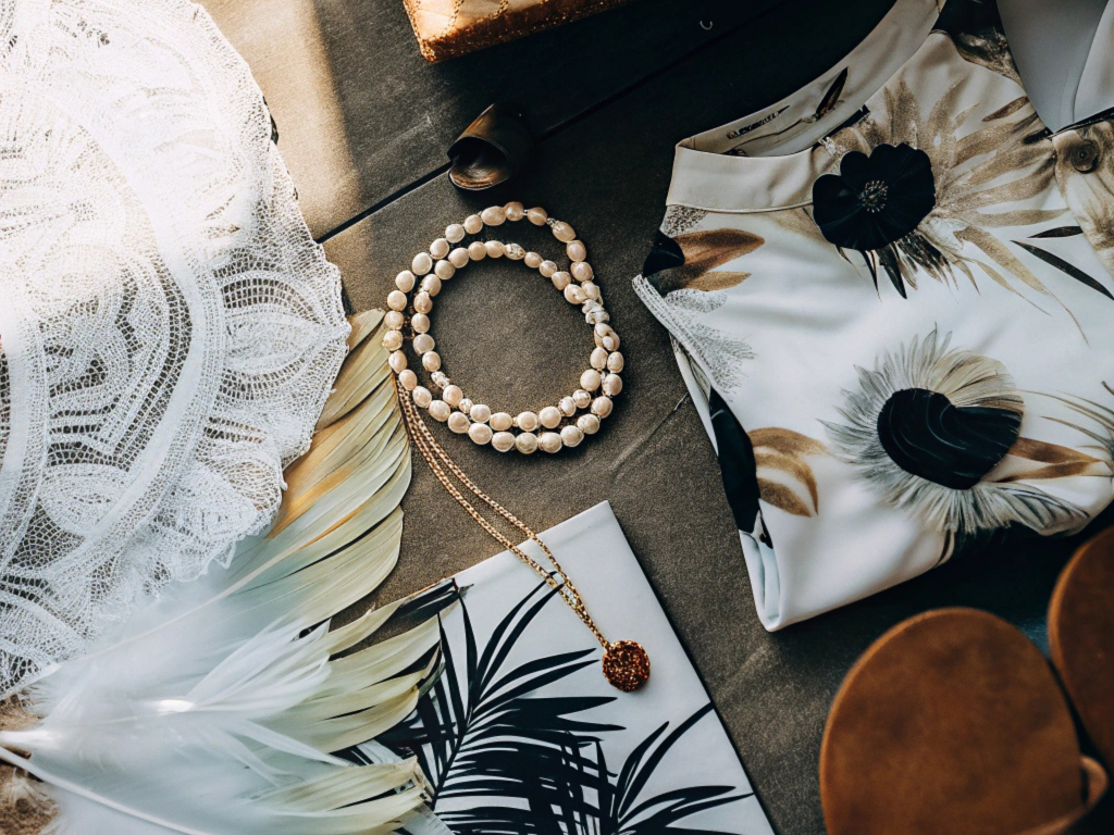 Elegant flat lay with floral dress, lace fabric, pearl necklace, feathers, and a woven clutch.