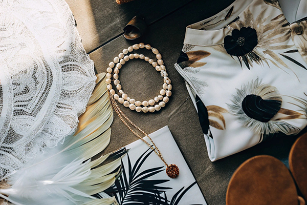 Elegant flat lay with floral dress, lace fabric, pearl necklace, feathers, and a woven clutch.