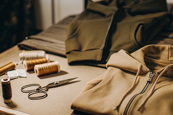Hoodies, spools of thread, scissors, and fabric on a wooden workspace.