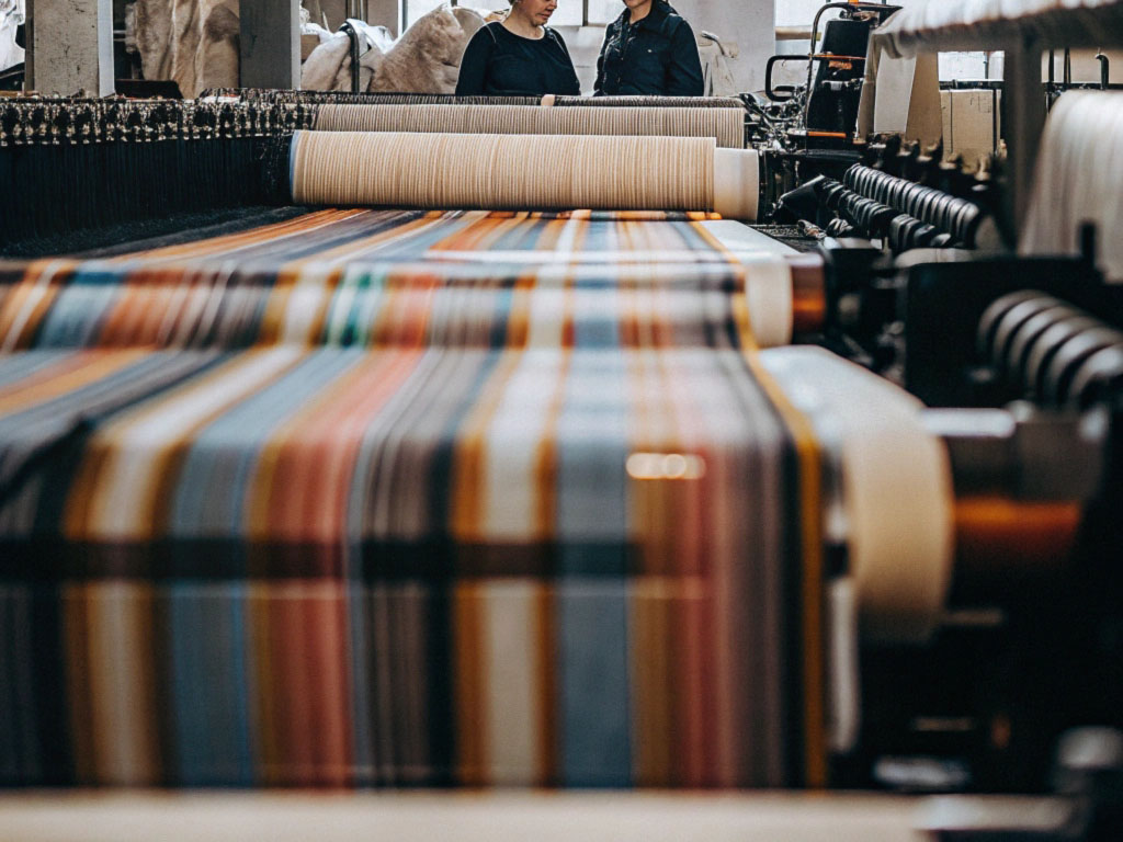 Colorful striped fabric being woven on an industrial loom in a textile factory with two workers in the background.