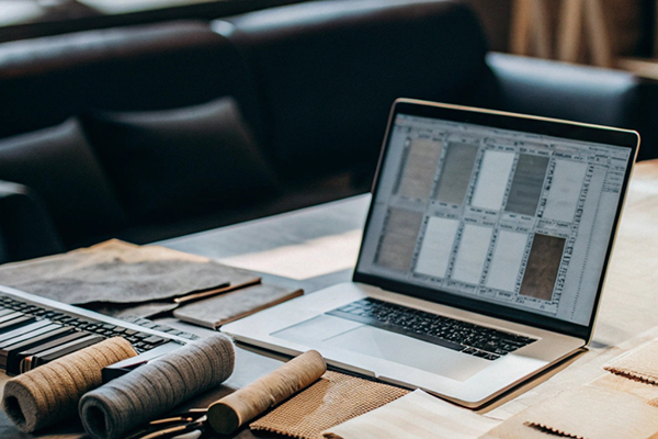 Laptop displaying fabric designs, with fabric samples, spools, and scissors on a worktable in a cozy studio.