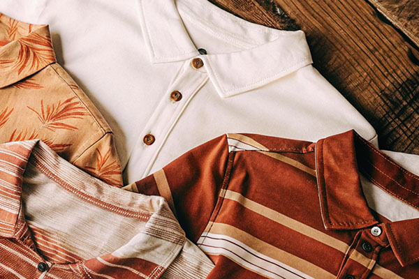 Four neatly arranged polo shirts in beige, white, and earthy red tones, featuring stripes, solid colors, and leaf patterns on a wooden surface.