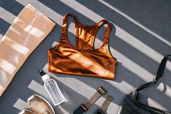 Flat lay of fitness items including a bronze sports bra, leggings, water bottle, smartwatch straps, sneakers, and a gym bag in sunlight.